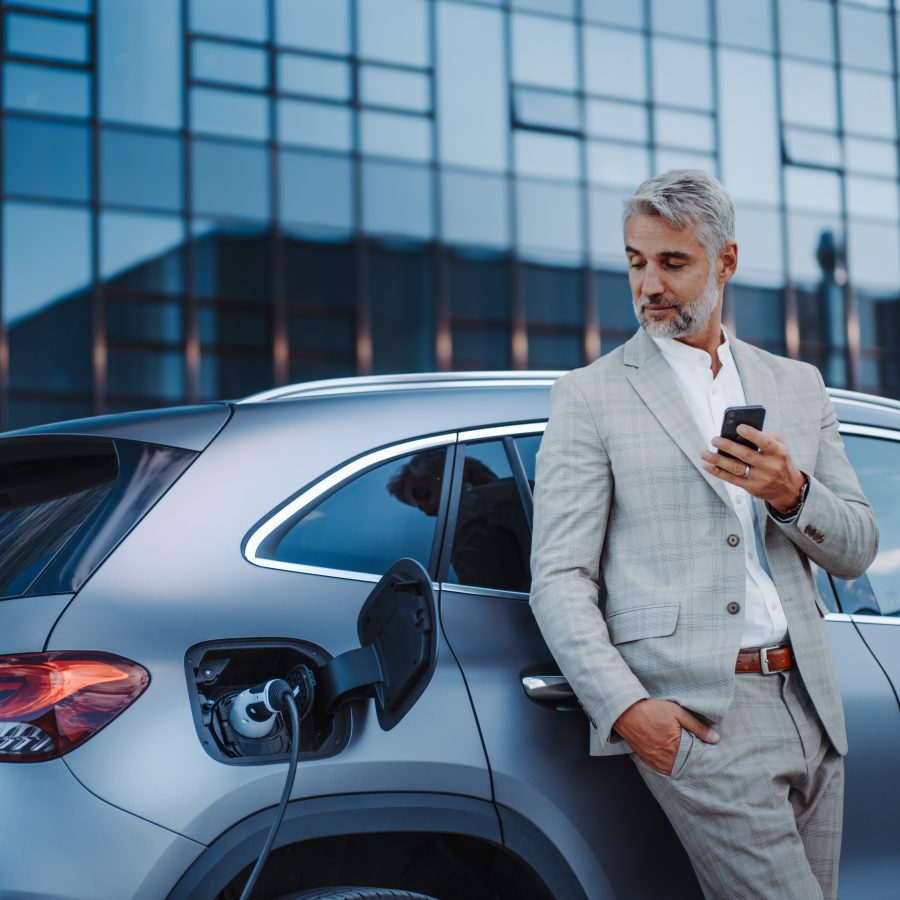 A businessman holding smartphone while charging car at electric vehicle charging station, closeup.